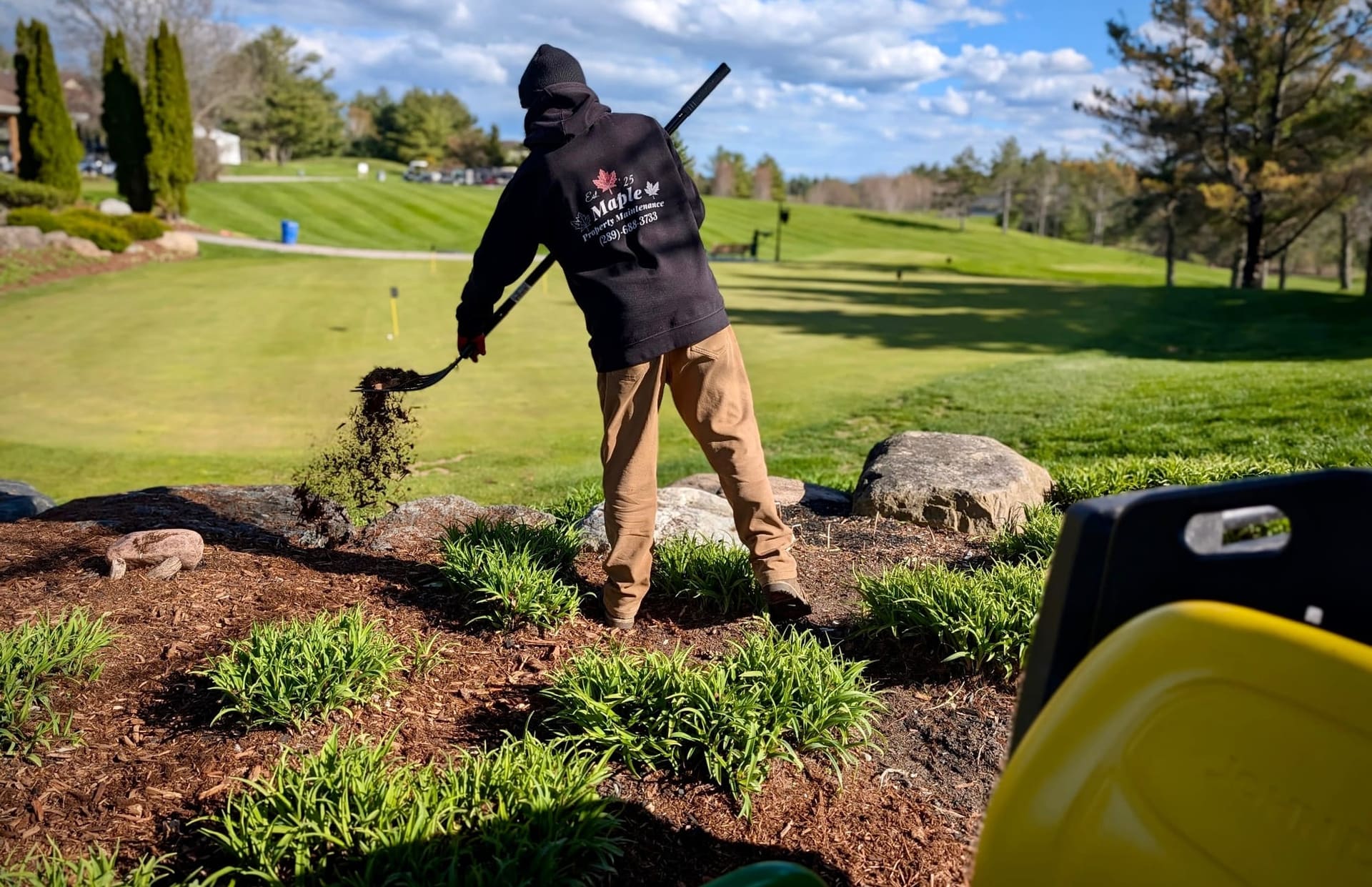 Crew member spreading fresh mulch with a pitchfork on a garden bed beside a golf course green